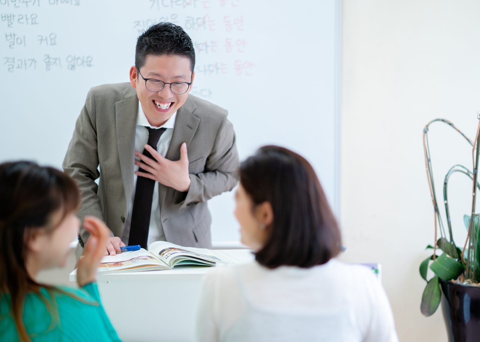 韓国語が書かれたホワイトボードの前で、開いた教材がある机をはさみ、スーツ姿の男性と座っている女性2人が話している様子